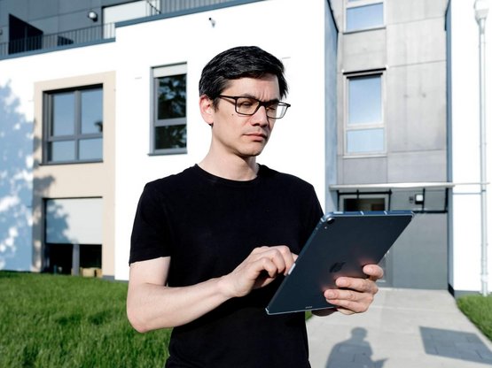 Ein junger Mann steht mit einem Tablet vor einer Reihe Einfamilienhäuser und tippt konzentiiert Daten in das Tablet ein.