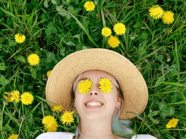 Eine junge Frau mit Strohhut liegt auf einer grünen Wiese. Auf Ihren Augen liegen zwei gelbe Blumen