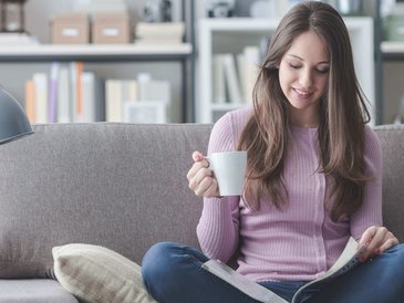 Eine junge Frau sitzt mit einer Tasse auf der Couch. Sie blättert in einem Magazin das auf Ihren Schoß liegt